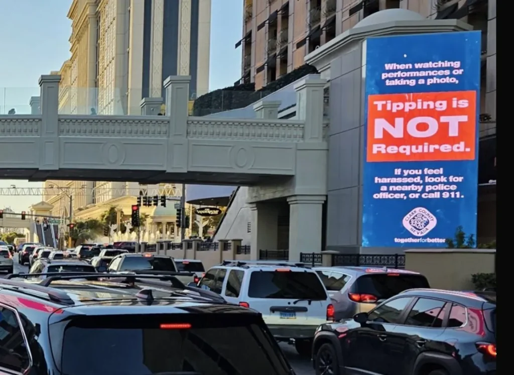 Cartel digital de Clark County en la Franja de Las Vegas advirtiendo que propinar a artistas callejeros no es obligatorio, con el letrero de bienvenida al fondo
