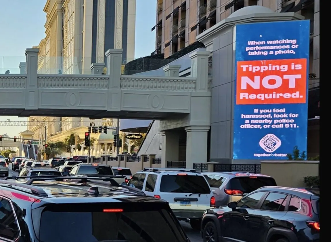 Cartel digital de Clark County en la Franja de Las Vegas advirtiendo que propinar a artistas callejeros no es obligatorio, con el letrero de bienvenida al fondo